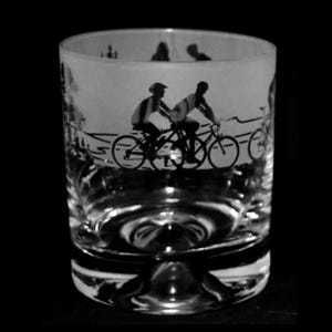 May include: A clear glass tumbler with a black and white illustration of cyclists on a road. The design wraps around the glass, featuring silhouettes of people riding bicycles against a landscape background. The glass has a thick base.