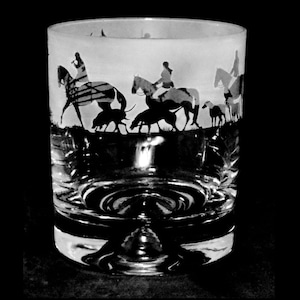May include: A clear, round drinking glass featuring a black and white silhouette design of riders on horseback, dogs, and a landscape. The glass has a thick base and a slightly flared rim, suitable for various beverages.