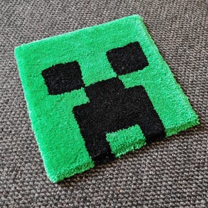 May include: A square, green rug with a black pixelated Creeper face design. The rug is made of a soft, textured material and is placed on a grey surface. The Creeper design is a popular character from the Minecraft video game.