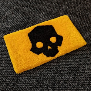 May include: A rectangular, yellow, tufted rug featuring a black skull design. The skull has two eye sockets and a nose cavity. The rug is placed on a dark grey, textured surface.