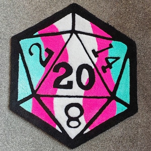 May include: A tufted rug shaped like a twenty-sided die, featuring a black border and black lines forming triangular segments. The surface displays alternating wavy stripes of bright pink, white, and teal, with the black numbers 20, 2, 14, and 8.