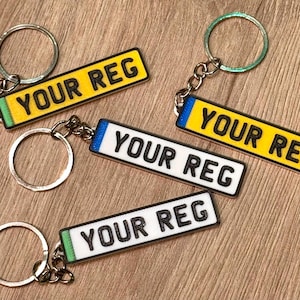 May include: Keyrings featuring miniature license plate designs. The rectangular keyrings display the text "YOUR REG" in black. The license plates have yellow, white, blue, and green borders. The keyrings are attached to silver key rings.