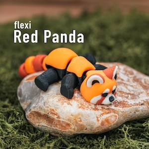 May include: A 3D-printed, orange and black flexi Red Panda toy rests on a stone, set against a backdrop of green moss. The text "flexi Red Panda" is displayed above the toy. The panda has a detailed design.