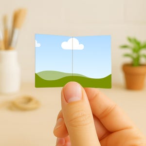 May include: A small, open card held by a hand, featuring a landscape illustration of a blue sky with a white cloud and green hills. The card is rectangular with a vertical crease down the middle.