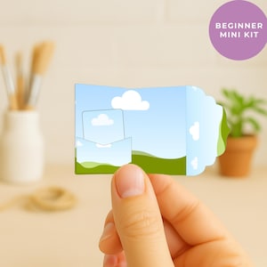 Puede incluir: Una pequeña tarjeta de papel plegada con un diseño de paisaje, sostenida por una persona. La tarjeta presenta un cielo azul, nubes blancas y colinas verdes. El texto "BEGINNER MINI KIT" está en un círculo morado.