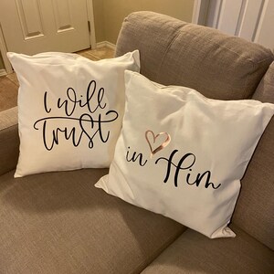 May include: Two cream-coloured throw cushions with black script. One cushion reads "I will trust" and the other reads "in Him" with a rose gold heart. The cushions are on a neutral-coloured sofa.