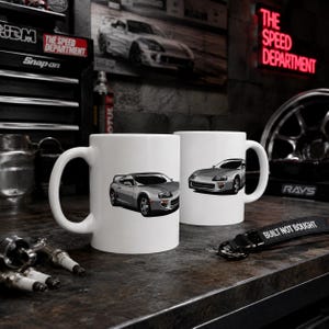 May include: Two white ceramic mugs featuring a graphic illustration of a silver sports car. One mug displays the car from a side angle, while the other shows the front view. The mugs are positioned on a dark surface alongside automotive parts.