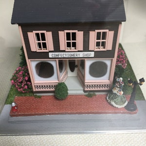 May include: A miniature confectionery shop with a pink facade and a sign that reads "Confectionery Shop". The shop has two windows with pink trim and a small brick walkway in front. There is a miniature person in a green dress standing in front of the shop.