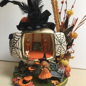 May include: A white pumpkin with a black and white spiderweb design is decorated with a black witch's hat and a miniature witch doll holding a pumpkin. The pumpkin is open to reveal a miniature room with a black and orange patterned rug, a small table, and two chairs. The pumpkin is surrounded by fall foliage and a small bench.