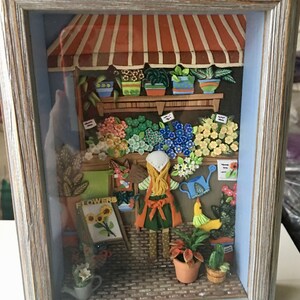 Puede incluir: Una escena de tienda de flores en miniatura con una niña con un vestido amarillo y un delantal rojo regando plantas. La tienda tiene un toldo a rayas rojas y blancas y un letrero que dice "FLOWERS".