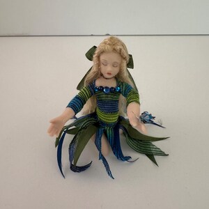 May include: A miniature doll with blonde hair and a green bow. The doll wears a blue, green, and black striped top with blue jewel accents. The doll's skirt is made of green and blue ribbons.