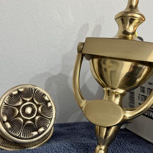 Puede incluir: Primer plano de dos herrajes de puerta de latón. Uno es un pomo redondo con un diseño floral, y el otro es un aldaba con un asa. Ambas piezas tienen un acabado dorado brillante.