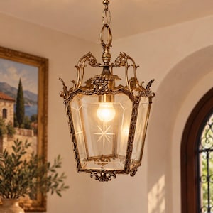 May include: A hanging lantern pendant light with an antique gold metal frame featuring ornate scrollwork and decorative accents. The fixture has four clear glass panels, each etched with a central starburst design, and a single light bulb inside.