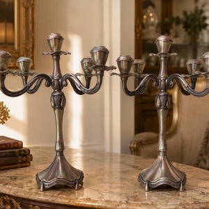 Puede incluir: Dos candelabros de metal plateado a juego, cada uno con un tallo central y cuatro brazos curvos que se extienden hacia afuera. Los candelabros tienen un acabado pulido, detalles florales decorativos en la base de los brazos y una base ancha y festoneada.