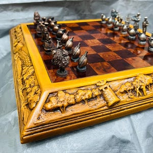 Puede incluir: Un juego de ajedrez detallado con un tablero de madera y piezas de metal intrincadamente diseñadas. El tablero presenta un patrón de vetas de madera oscura, y el marco está tallado con una escena de paisaje. Las piezas de ajedrez son en tonos bronce y plata.
