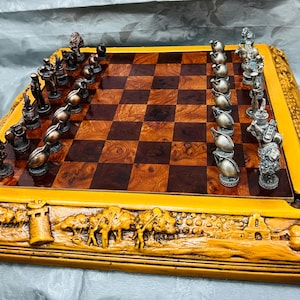 Pode incluir: Um conjunto de xadrez detalhado com um tabuleiro de madeira com um padrão xadrez em quadrados castanhos e castanho escuro. As peças de xadrez são metálicas, com uma borda detalhada esculpida com uma cena desértica.