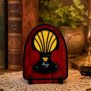 Könnte beinhalten: Ein Radio im Vintage-Stil mit roter und schwarzer Oberfläche und gelbem Lautsprechergrill. Das Radio hat ein Zifferblatt und zwei Knöpfe und steht auf einer Spitzendecke. Im Hintergrund sind antike Bücher und eine Lampe zu sehen.