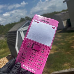 May include: A pink Nokia flip phone with a keypad and screen. The phone has the text "NOKIA" at the top and "The Glitch Lab" at the bottom. The phone is held against a background of a blue sky and houses.