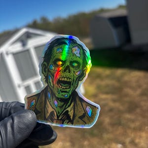 May include: Holographic sticker featuring a zombie illustration. The zombie has green skin, exposed teeth, and a decaying appearance. It is wearing a brown jacket and a shirt. The sticker has a rainbow-like, iridescent effect.