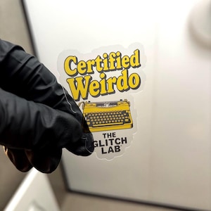 May include: A clear sticker with the text "Certified Weirdo" in yellow, above a yellow typewriter graphic. Below the typewriter is the text "THE GLITCH LAB". The sticker is held by a gloved hand.