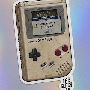 May include: A sticker featuring a vintage Nintendo Game Boy with a cracked screen displaying an error message that reads "damm, we're getting old." The Game Boy has a beige color with black and red buttons. The sticker also includes the text "TAL GLITCH LAB."