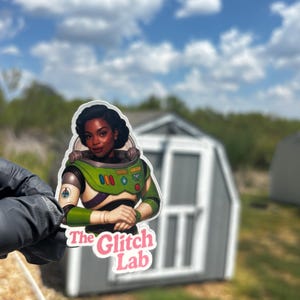 May include: A sticker featuring a woman in a green and white space suit, with the text "The Glitch Lab" in pink and white. The sticker is held up against a backdrop of a cloudy blue sky and a gray shed.