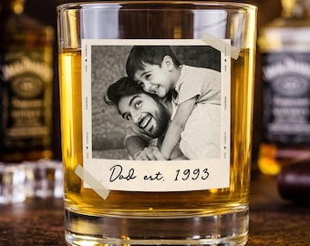 May include: A rocks glass filled with amber liquid, featuring a black and white photo of a man and child. The photo is taped to the glass and has the text "Dad est. 1993". Whiskey bottles are in the background.