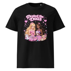 May include: Black t-shirt featuring a graphic of two cartoon women with long hair, surrounded by pink flowers, hearts, and stars. The text "Protect the Dolls" is above the women. A pink car, unicorn, and teddy bears are also in the graphic.