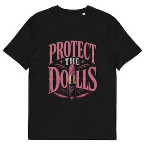 以下が含まれることがあります： 黒いTシャツ。ピンク色のグラフィックで「PROTECT THE DOLLS」の文字と口紅のイラストが描かれています。口紅は金色のチューブとハートのディテール。デザインは少し擦り切れたように見えます。