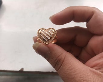 May include: A rose gold heart-shaped ring with a band of small, clear stones around the heart's edge. The heart is filled with rectangular, clear stones. The ring is held up by a hand.