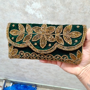 Pode incluir: Uma clutch de veludo verde com desenhos florais dourados com contas. A clutch apresenta um fecho de aba e é adornada com padrões intrincados de contas douradas, criando um acessório luxuoso e elegante. O design geral é simétrico e detalhado.