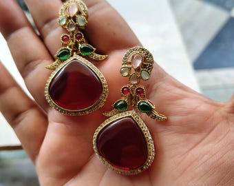 May include: A pair of gold-toned drop earrings. Each earring features a large, teardrop-shaped, deep red stone surrounded by small, clear stones. The earrings have accents of green, pink, and white stones.