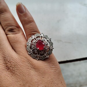 May include: A silver-toned ring featuring a large, oval-cut red gemstone at its center, surrounded by a detailed, floral-inspired design. The ring has a cluster of smaller, clear stones and intricate metalwork, creating a bold and elegant statement piece.