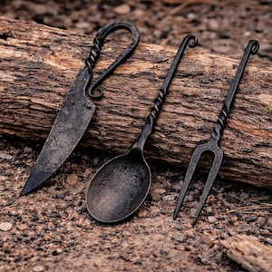 May include: Hand-forged black iron cutlery set including a knife, spoon, and fork. The handles have a twisted design, and the set rests on a weathered log. The knife blade is approximately 6 inches long.