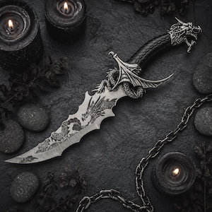 May include: A silver-toned dagger featuring a dragon-shaped pommel and a crossguard with dragon wing details. The blade displays an intricate etched dragon design. The handle is black with a textured grip, complemented by a metallic chain accessory.