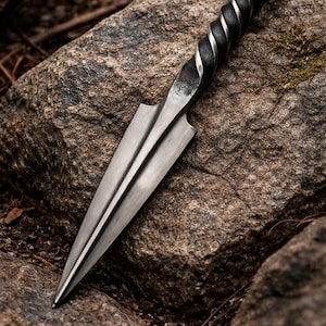 Peut inclure: Un poignard en acier forg&eacute; &agrave; la main, avec un manche torsad&eacute; et une lame &agrave; double tranchant. Le poignard est pr&eacute;sent&eacute; sur une grosse pierre textur&eacute;e, mettant en valeur son savoir-faire. La lame mesure environ 25 cm.