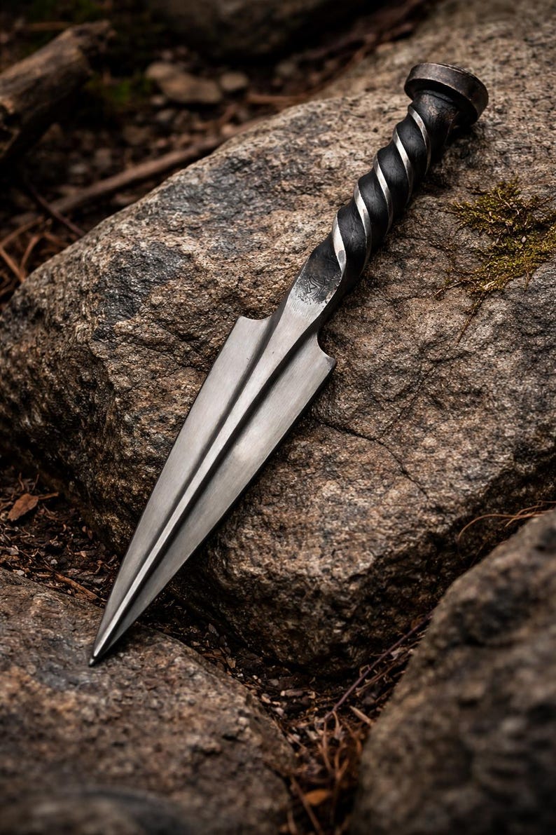 Peut inclure: Un poignard en acier forg&eacute; &agrave; la main, avec un manche torsad&eacute; et une lame &agrave; double tranchant. Le poignard est pr&eacute;sent&eacute; sur une grosse pierre textur&eacute;e, mettant en valeur son savoir-faire. La lame mesure environ 25 cm.