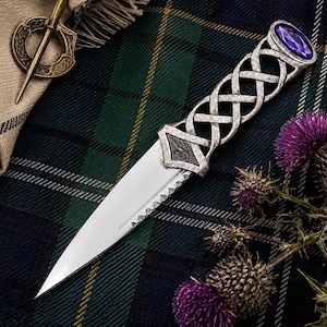 May include: A silver-toned metal sgian-dubh knife featuring an intricate Celtic knotwork handle, a pointed steel blade, and a purple oval gemstone set at the pommel. The knife is displayed alongside a bronze-colored circular brooch pin.