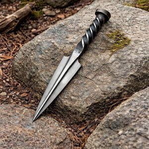 Peut inclure: Un poignard en m&eacute;tal forg&eacute; &agrave; la main avec un manche torsad&eacute; et une lame pointue et tranchante. Le poignard est de couleur argent&eacute;e et repose sur un gros rocher gris. La poign&eacute;e a un aspect sombre et textur&eacute;. L'arri&egrave;re-plan pr&eacute;sente d'autres rochers et du feuillage.