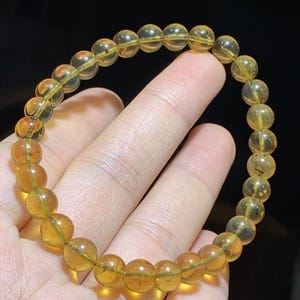 May include: A bracelet made of translucent, golden-yellow beads. The spherical beads are strung together, forming a circular shape. The bracelet is held against a dark background, highlighting the colour and clarity of the beads.