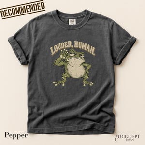 Puede incluir: Una camiseta de manga corta gris carbón con un gráfico de una rana verde con una mano cerca de la oreja. Sobre la rana, el texto LOUDER, HUMAN aparece en color beige. La prenda tiene cuello redondo y una etiqueta de la marca Comfort Colors.