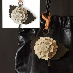 Peut inclure: Un charm de sac en cuir en forme de fleur d'hortensia. Il présente des pétales blanc cassé avec de petits détails bruns, une feuille vert foncé et un anneau doré. Le charm est attaché à un sac en cuir noir.