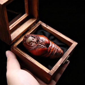 Peut inclure: Une boîte en bois avec un couvercle à charnière, contenant une sculpture sculptée d'un bébé endormi enveloppé dans un cocon rouge. Le visage du bébé est visible, avec les yeux fermés. La boîte est tenue dans une main.