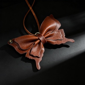 May include: A brown leather butterfly-shaped crossbody bag with a long strap. The bag features detailed wing patterns and a central band. The bag is made of textured leather and has a small metal clasp on the side.