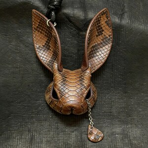 May include: A brown leather rabbit pendant with a snake-skin pattern. The pendant has long ears and a small chain with a teardrop-shaped charm. The pendant is attached to a black leather cord.