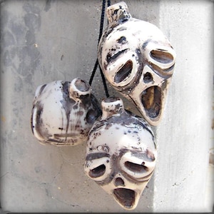 May include: Three off-white ceramic face pendants with dark brown markings hang against a grey background. Each pendant has a unique, expressive face with open mouths and eye sockets. The pendants are strung on a black cord.
