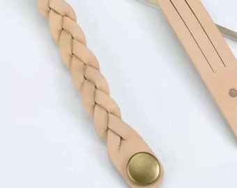May include: Close-up of a light tan leather braided strap with a gold-colored button. The strap is part of a larger object, possibly a handbag or accessory. The background is a light neutral color.