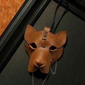 May include: A brown leather mask in the shape of a cat's face, with stitched details and eye holes. The mask has a black cord for hanging and a silver chain attached to the bottom. The mask is set against a dark background.