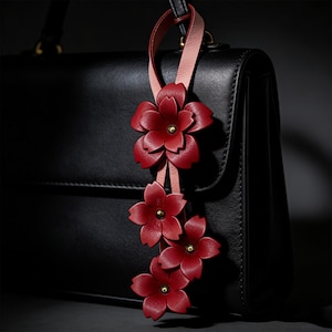 Puede incluir: Un adorno de bolso de cuero con tres flores rojas y centros dorados. Las flores están unidas a una correa de cuero rosa y roja, colgando de un bolso de mano de cuero negro. El adorno añade un toque decorativo al bolso.