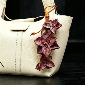 May include: Cream-colored leather tote bag with a decorative burgundy leather flower charm. The charm features three dimensional flower shapes with detailed veining, attached with a brown leather cord.